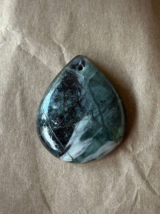 A polished teardrop-shaped gemstone pendant with a dark green, black, and white marbled pattern, resting on a textured brown paper background.