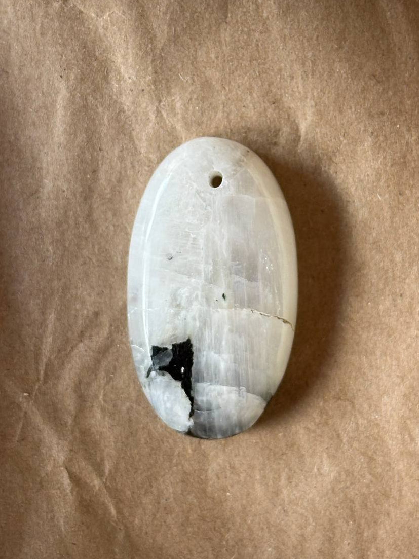 An oval polished stone pendant with a natural fissure and dark inclusions, resting on a textured brown paper background.