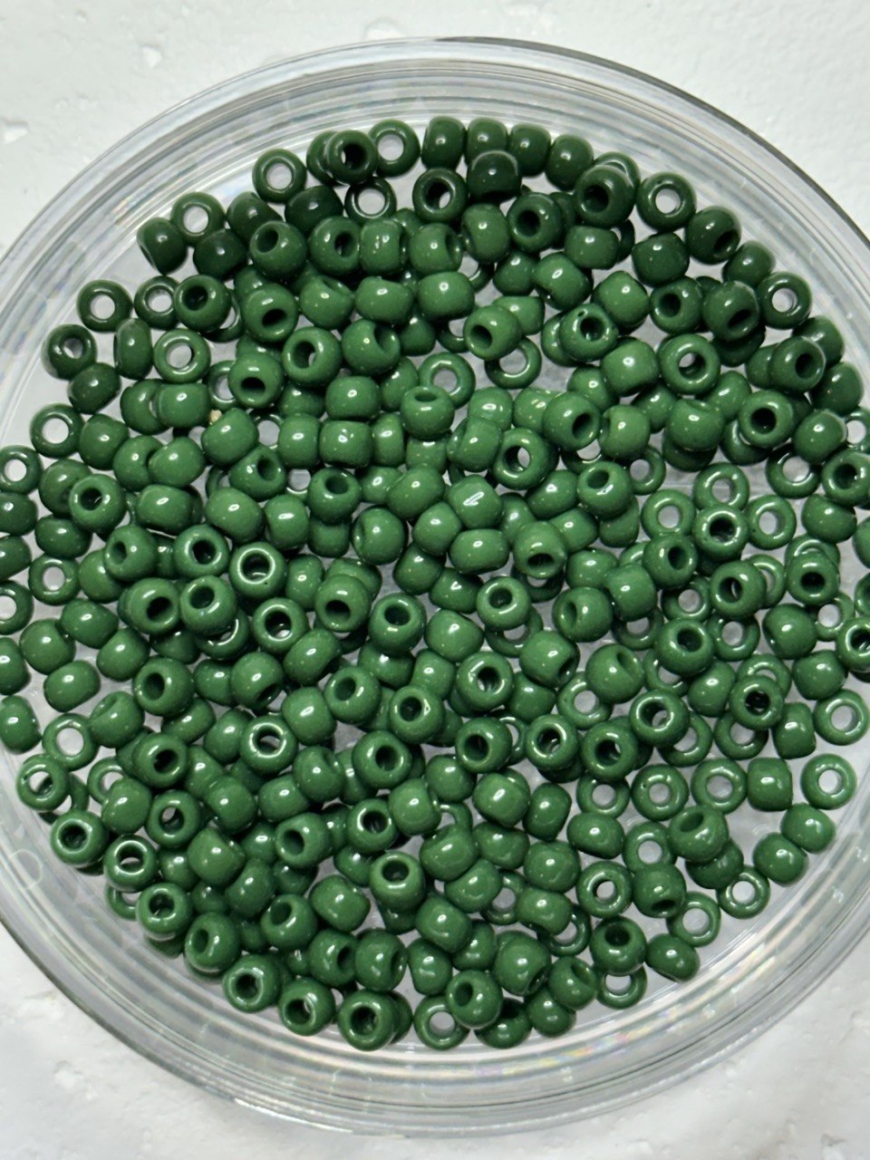 Green seed beads with a glossy finish, piled inside a clear glass petri dish on a light textured background.