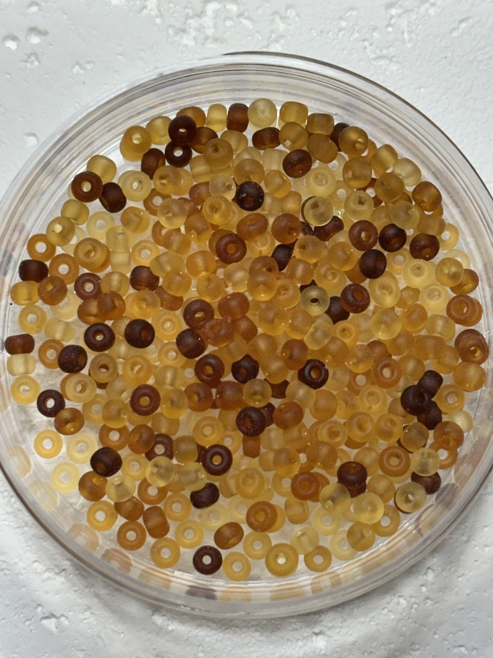 Small translucent ring-shaped beads in amber, yellow, and dark brown tones, photographed from above inside a clear round dish on a white textured surface.