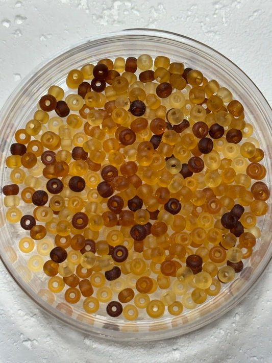 Small translucent ring-shaped beads in amber, yellow, and dark brown tones, photographed from above inside a clear round dish on a white textured surface.