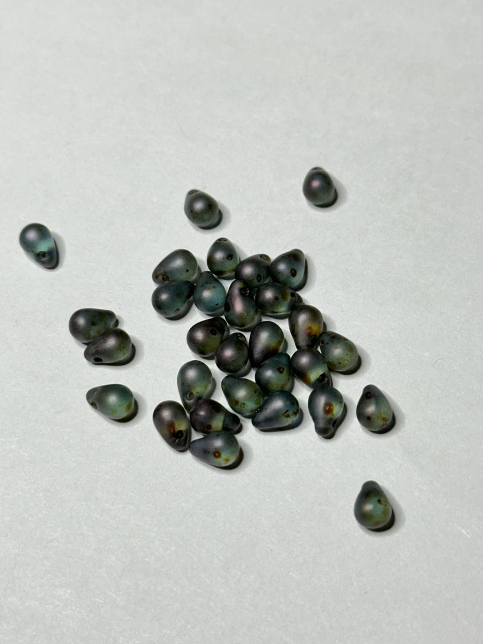 A small pile of dark iridescent teardrop-shaped beads scattered on a light gray background.