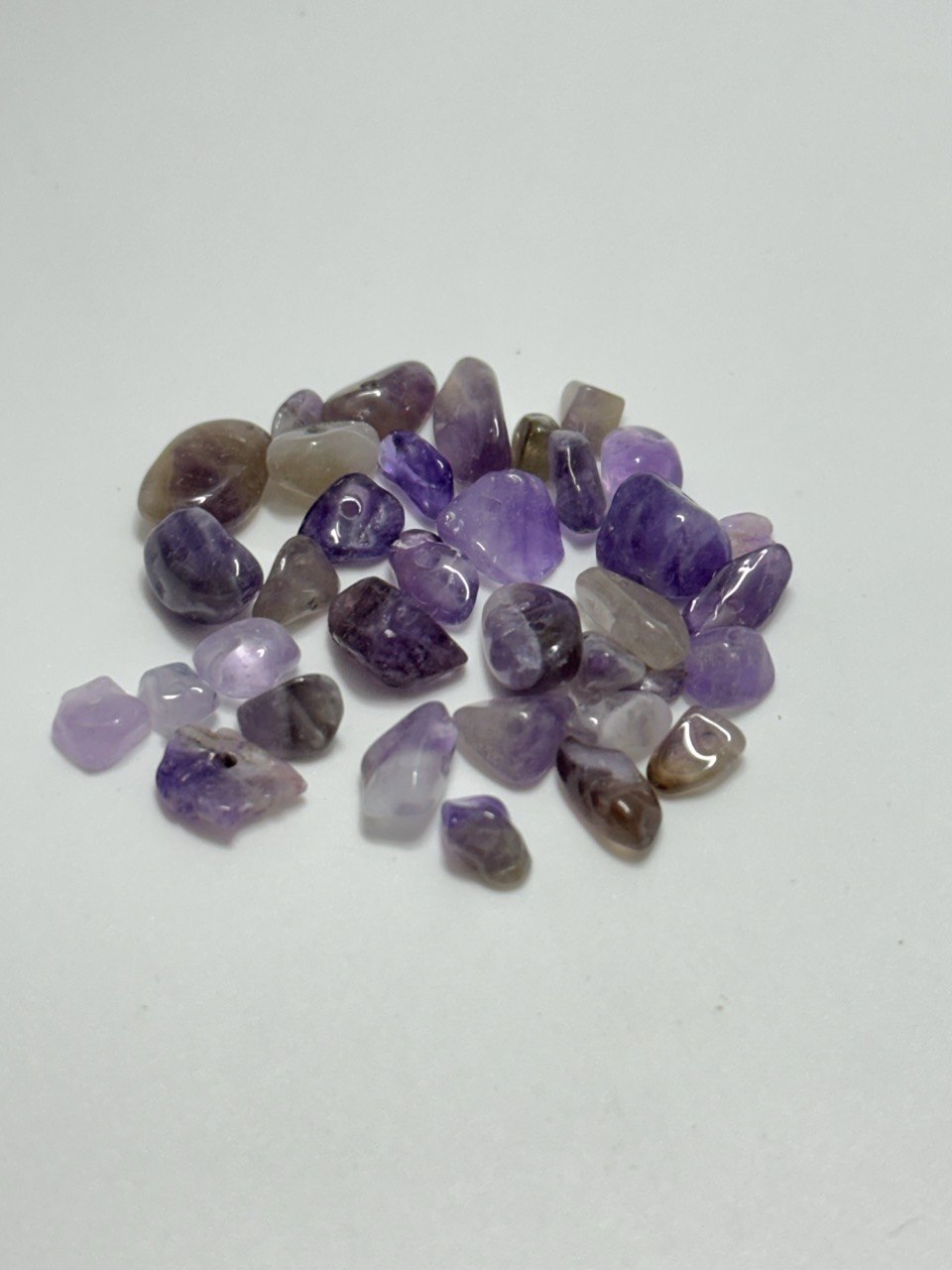 A small pile of polished tumbled purple stones, including amethyst and smoky quartz varieties, arranged on a plain white surface.