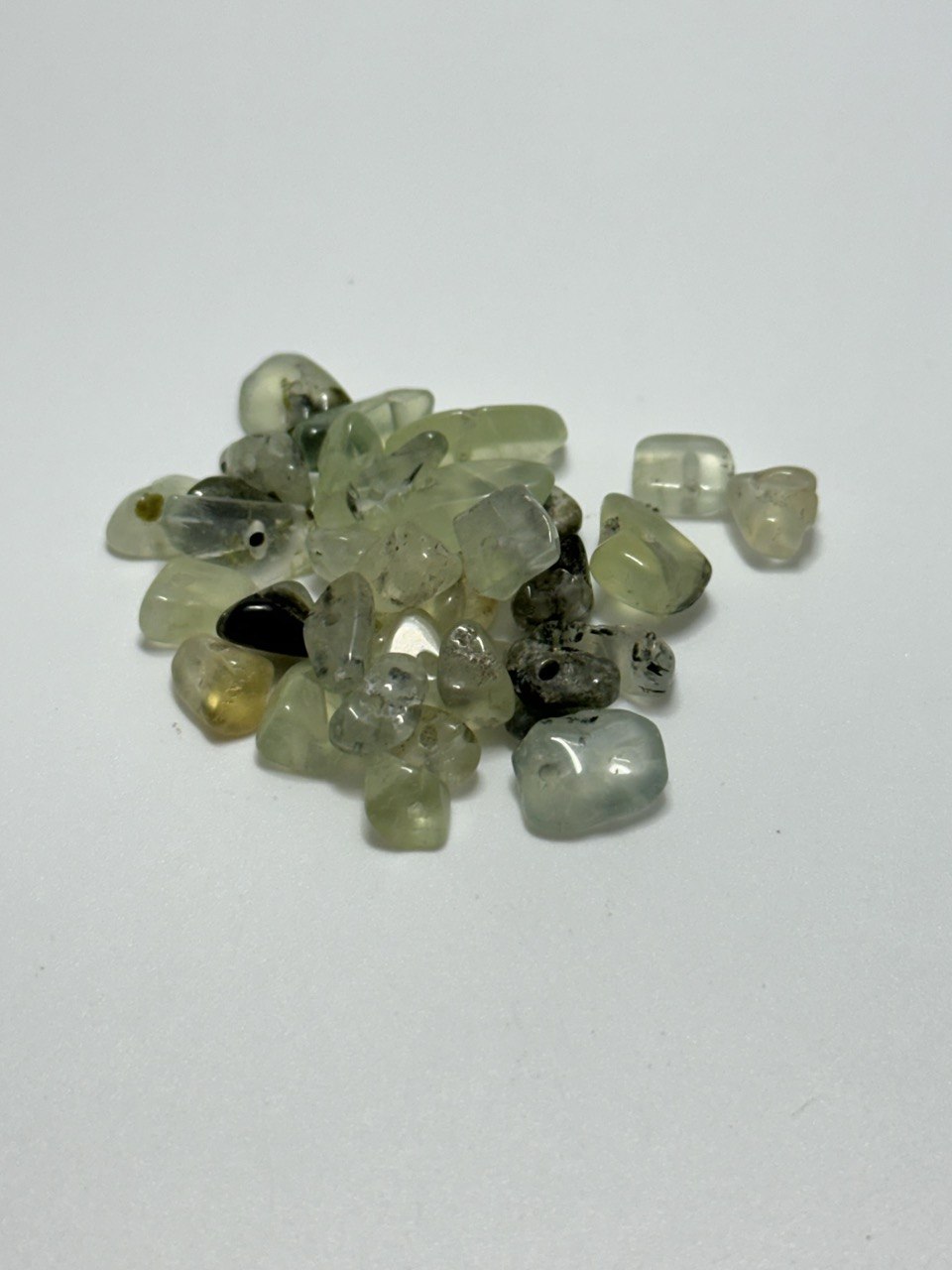 A small pile of polished tumbled gemstones in various translucent shades of green, gray, and clear, arranged on a plain white surface.