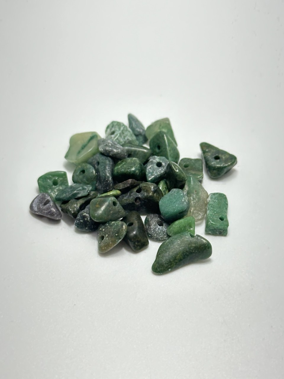 A pile of small green stone beads with irregular shapes and pre-drilled holes, photographed on a plain white background.