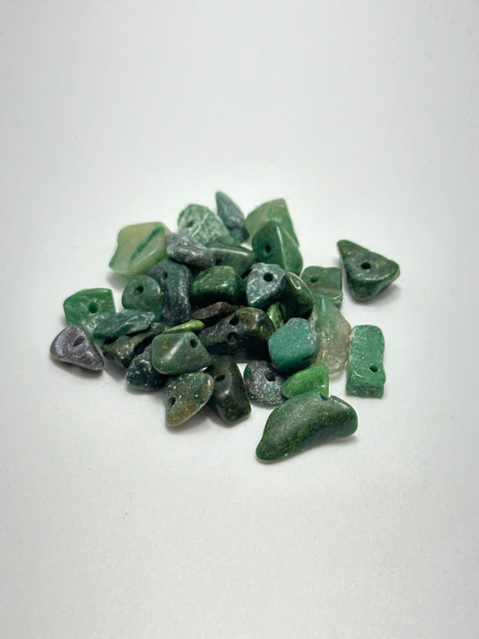 A pile of small green stone beads with irregular shapes and pre-drilled holes, photographed on a plain white background.