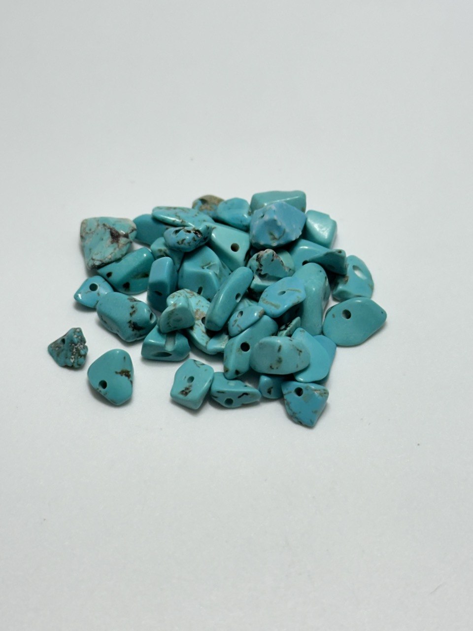 A pile of small turquoise stone beads with holes, showing natural matrix inclusions, on a plain light gray background.