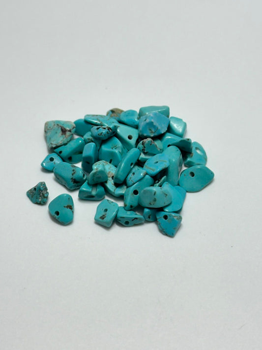 A pile of small turquoise stone beads with holes, showing natural matrix inclusions, on a plain light gray background.