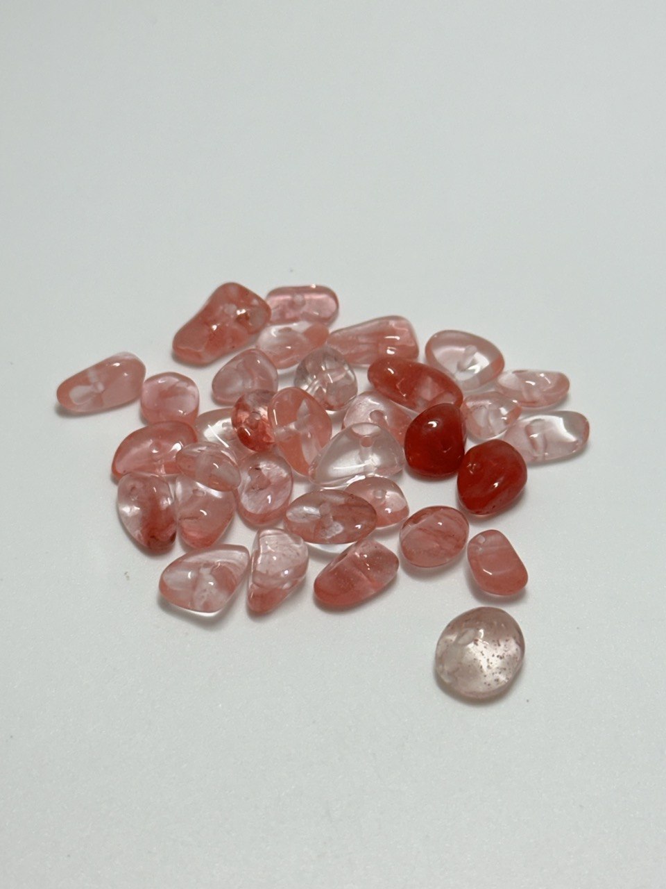 A small pile of polished tumbled gemstones in soft pink and translucent red tones, with one slightly larger pale pink stone placed below the pile, photographed on a plain light background.