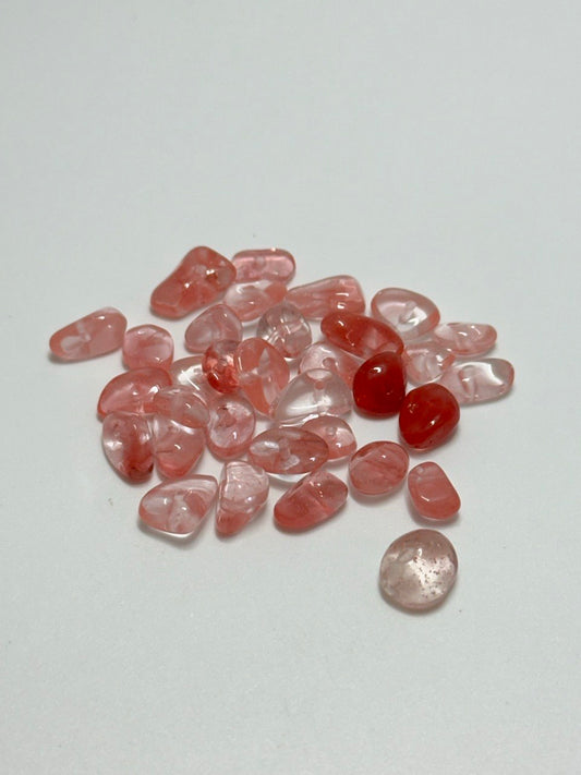 A small pile of polished tumbled gemstones in soft pink and translucent red tones, with one slightly larger pale pink stone placed below the pile, photographed on a plain light background.