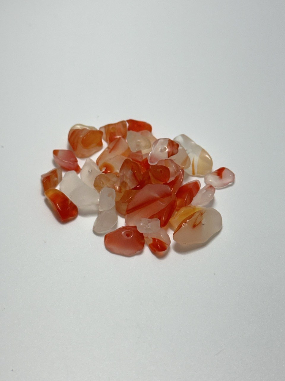 A small pile of polished tumbled carnelian stone chips in shades of orange, red, and translucent white, arranged on a plain light background.
