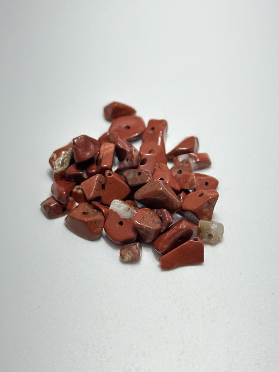 A small pile of red jasper stone beads with irregular, chunky shapes and pre-drilled holes, set against a plain light gray background.