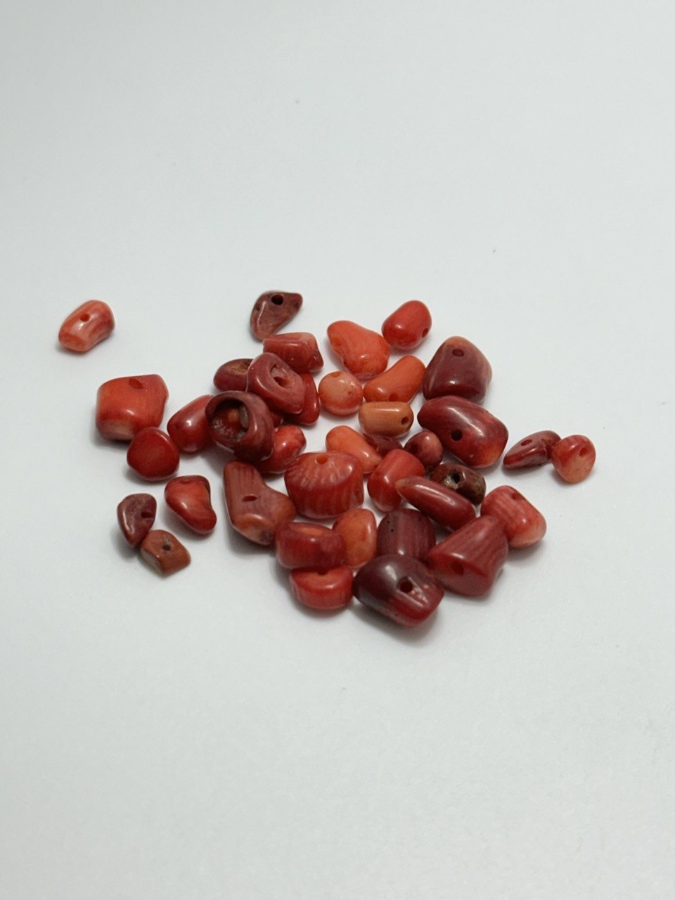 A pile of small polished red coral beads with varying shapes and sizes, arranged on a plain light gray background.