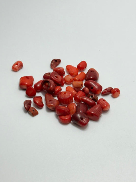 A pile of small polished red coral beads with varying shapes and sizes, arranged on a plain light gray background.