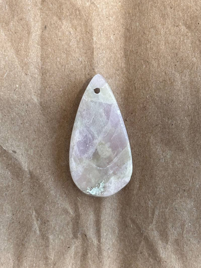 A teardrop-shaped polished gemstone pendant with natural lavender and white marbling, featuring a small hole at the top for hanging, displayed on a textured brown paper background.