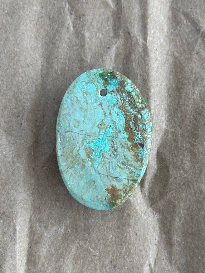 An oval chrysocolla cabochon stone with natural brown matrix and a small hole near the top, displayed on a textured gray background.