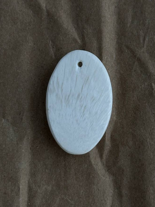 A small oval white stone pendant with a pre-drilled hole at the top, resting on a textured brown paper background.