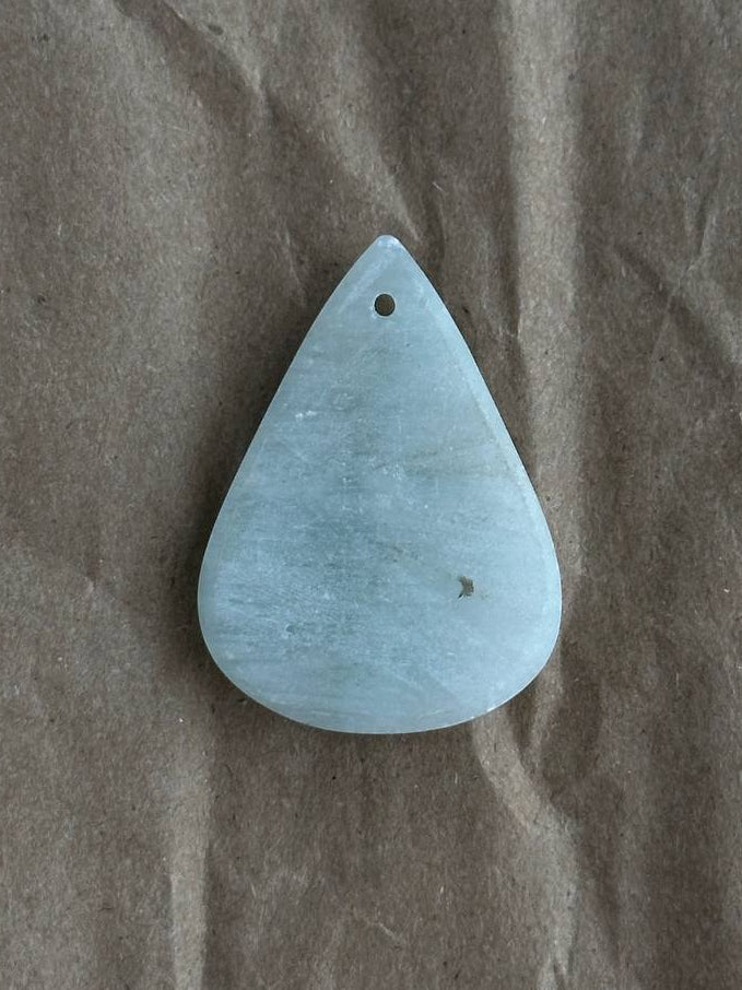 A teardrop-shaped light blue stone pendant with a small hole at the top, placed on a textured brown paper background.