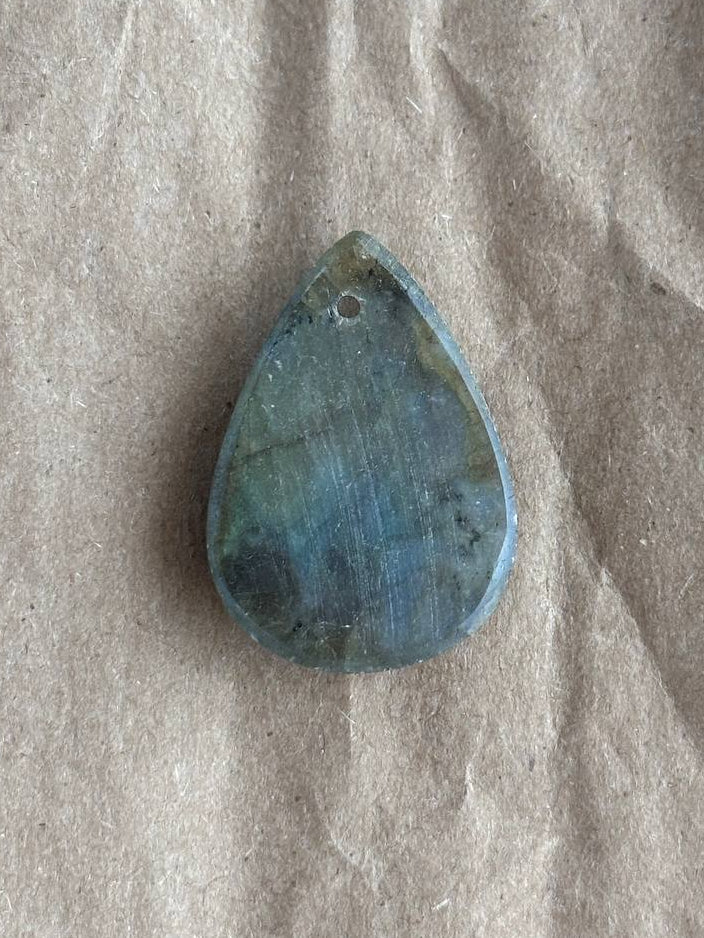 A teardrop-shaped labradorite gemstone pendant with a small hole at the top, resting on a textured beige paper background.