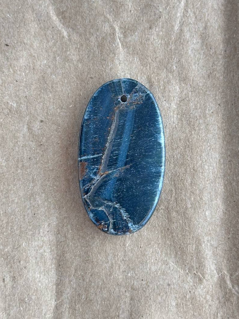 An oval blue stone pendant with natural white and gray veining, featuring a small hole at the top, displayed on a textured beige surface.