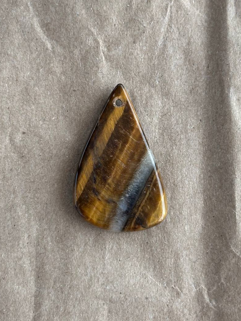 A teardrop-shaped polished gemstone pendant with natural tiger's eye bands of brown, gold, and blue, resting on a textured gray paper background.