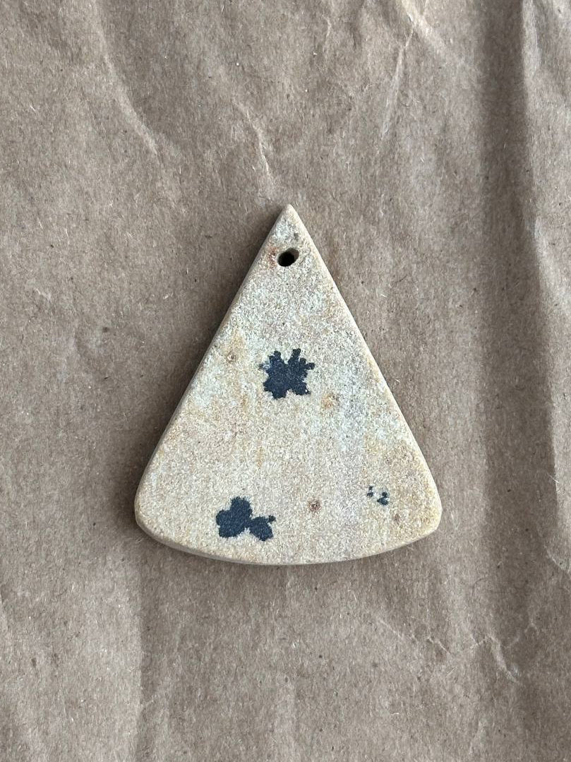 A triangular stone pendant with a pre-drilled hole at the top, featuring a speckled beige and white texture with dark blue floral inclusions, displayed on a textured brown paper background.
