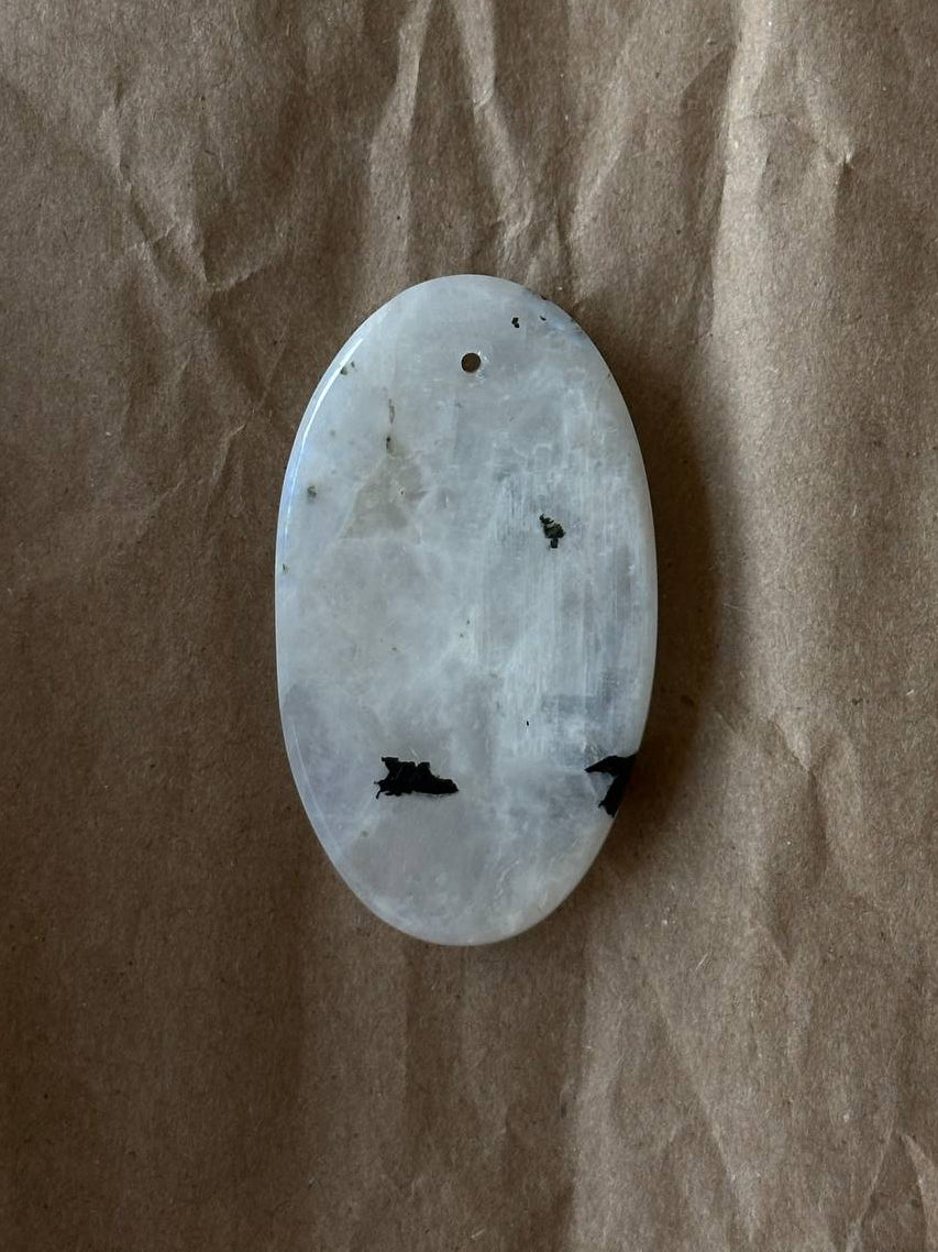 An oval-shaped polished stone cabochon with a white and gray marbled pattern and small black inclusions, resting on a brown textured paper background.