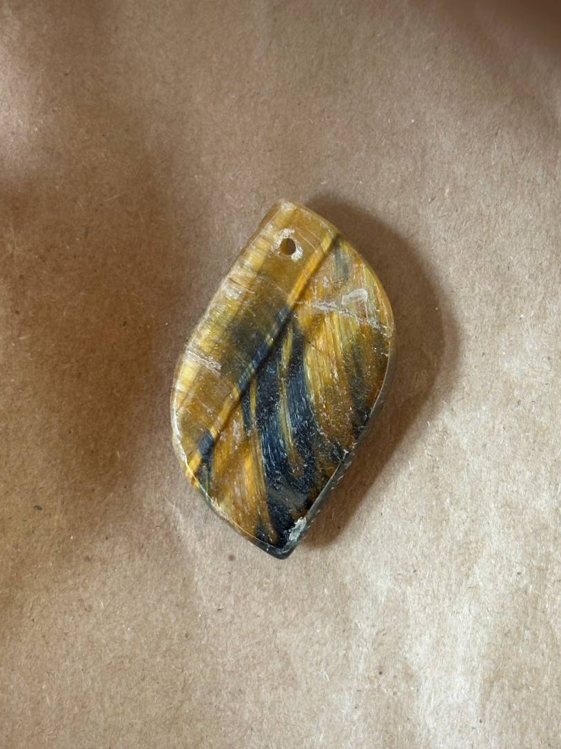 A small irregular-shaped pendant or charm with a hole at the top, featuring a marbled pattern of yellow, black, and white, resting on a brown textured surface.