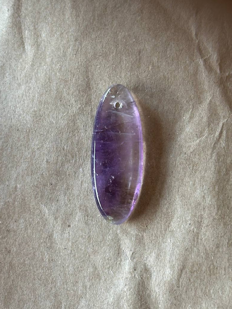 A polished oval amethyst gemstone pendant with a small hole at the top, resting on a textured beige paper background.
