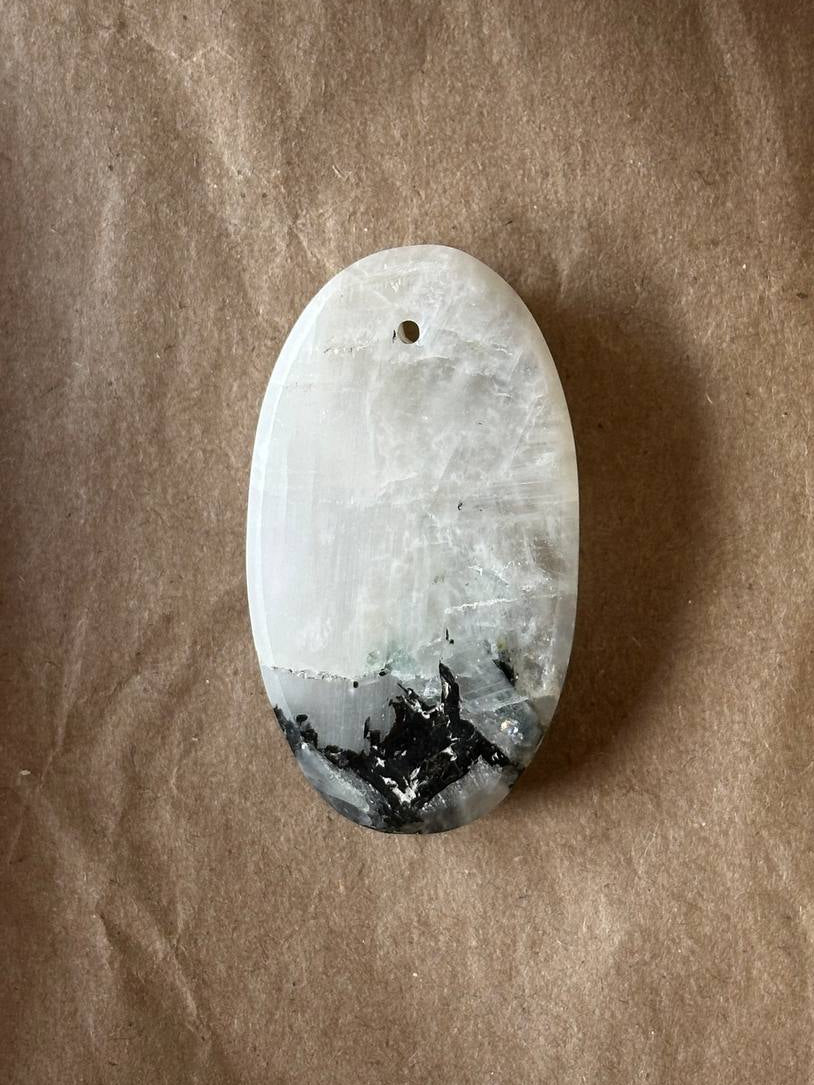 An oval-shaped polished stone pendant with a natural marbled pattern, featuring a white and gray upper section and a black and gray lower section, resting on a brown textured paper background.