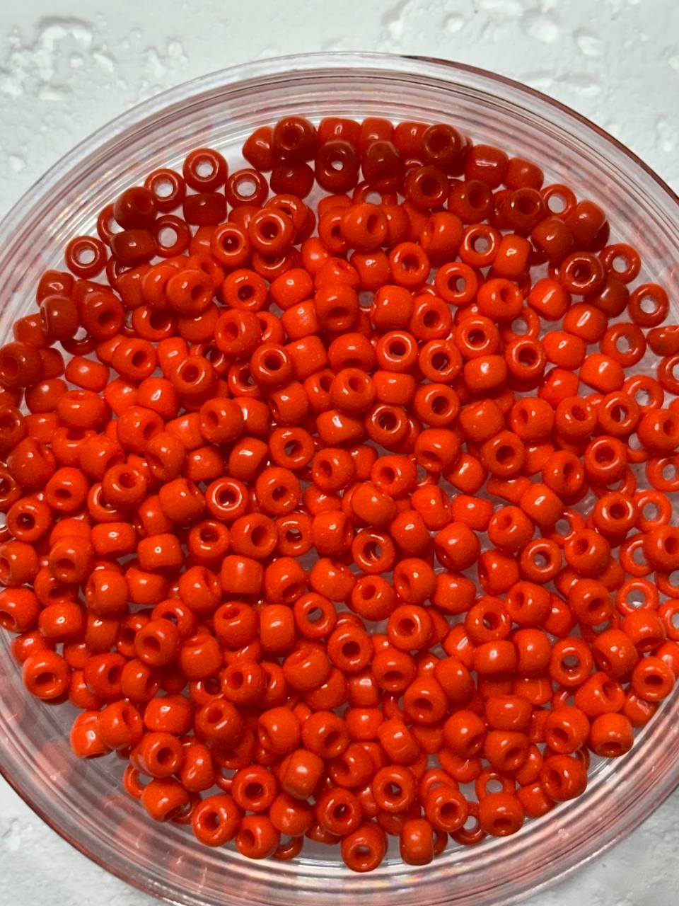 Bright orange-red round craft beads with hollow centers, piled inside a clear round plastic container on a light textured background.