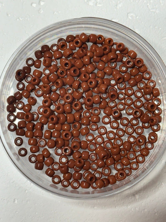 Brown seed beads with a glossy finish, arranged inside a clear plastic round container on a white textured surface.