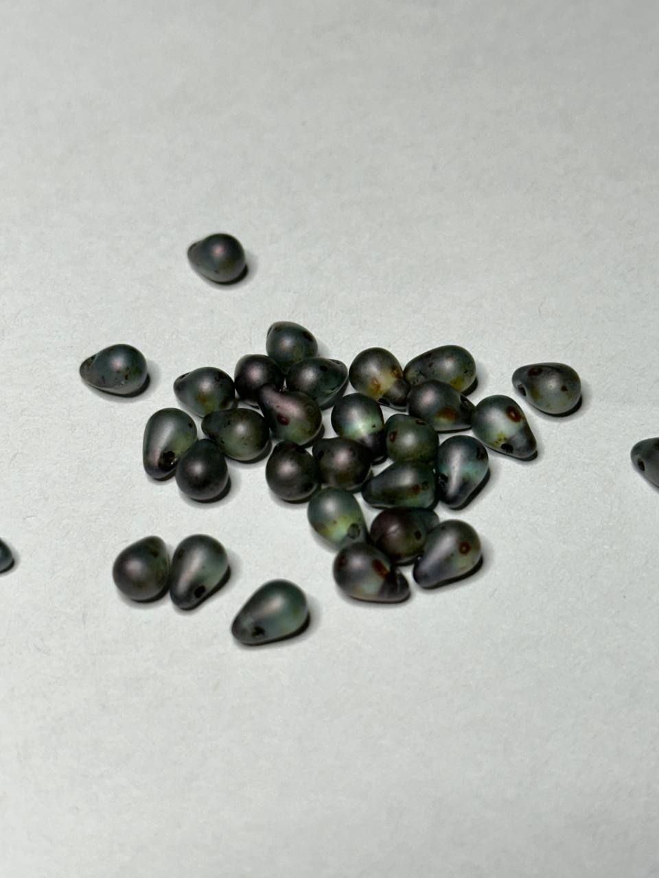A small pile of dark teardrop-shaped beads with a glossy, semi-matte finish and a central hole, scattered on a light gray textured background.