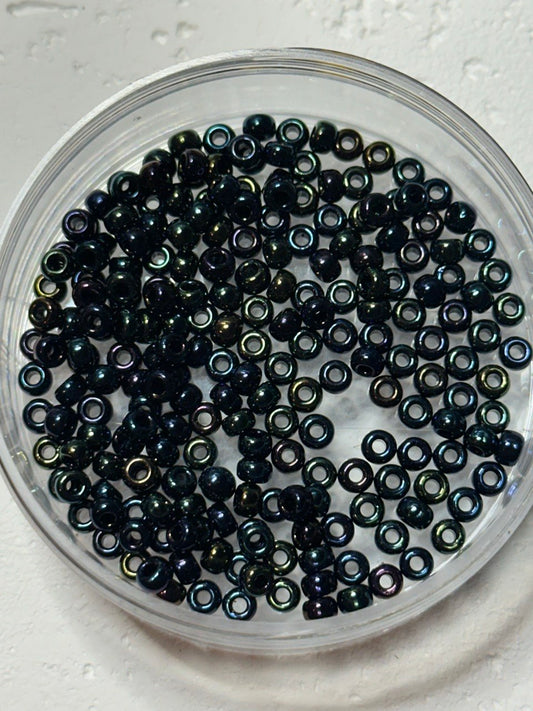 A top-down view of small iridescent dark beads inside a clear round plastic container, set on a light textured surface.