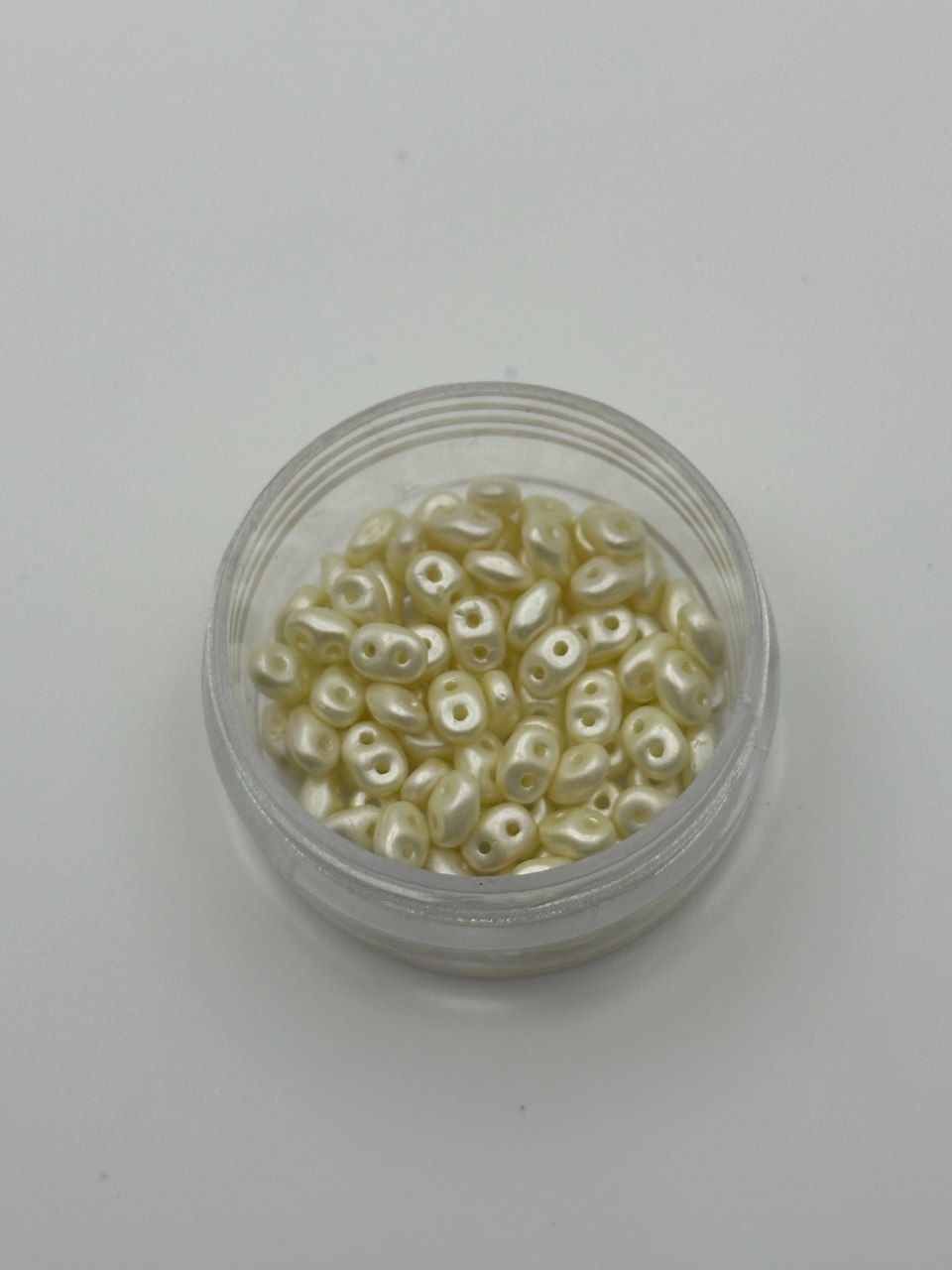 Small container filled with small light cream superduo beads on a light gray background