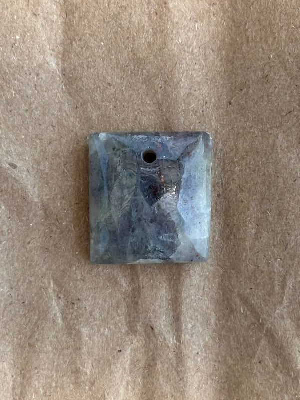 A square gray and blue stone pendant with a small hole at the top, resting on a textured brown paper background.