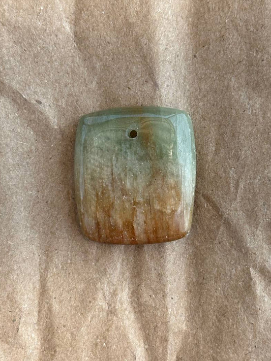 A rectangle-shaped polished stone pendant with rounded corners and a small hole at the top, featuring a color gradient from light green at the top to warm brown at the bottom, resting on a textured brown paper background.