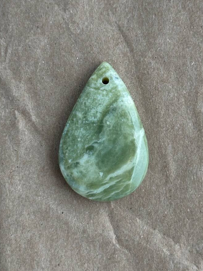 A polished green teardrop-shaped stone pendant with a small hole at the top, resting on a textured brown paper background.