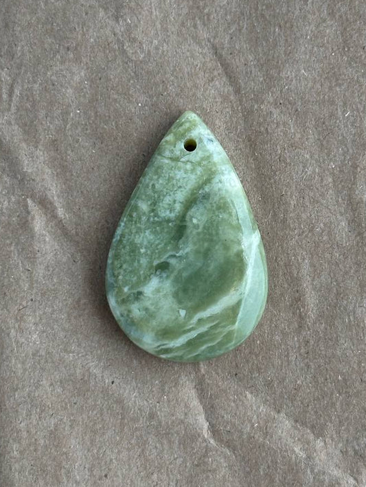 A polished green teardrop-shaped stone pendant with a small hole at the top, resting on a textured brown paper background.