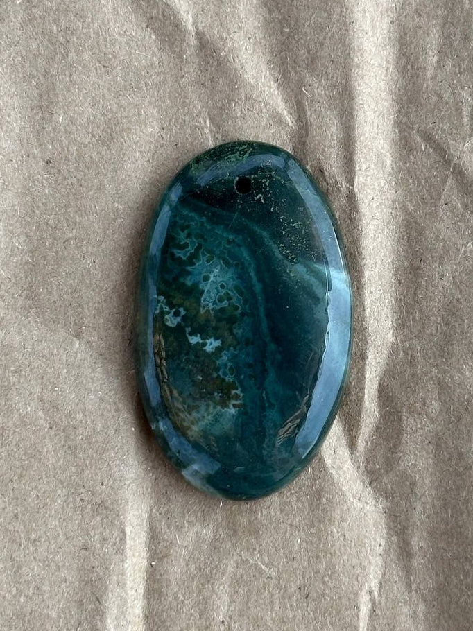 A polished oval cabochon gemstone with a marbled pattern of deep teal, blue, and green, resting on a textured beige paper background.