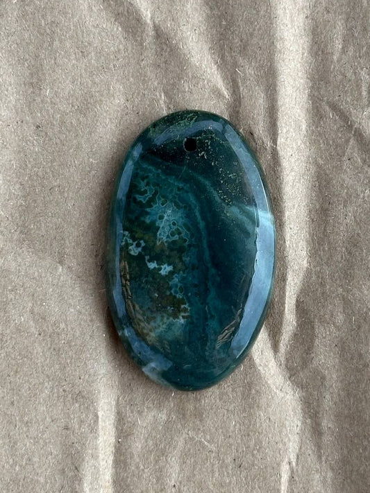A polished oval cabochon gemstone with a marbled pattern of deep teal, blue, and green, resting on a textured beige paper background.