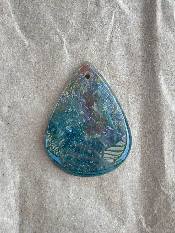 A teardrop-shaped moss agate pendant with a swirling blue, green, and purple marbled pattern, resting on a textured beige paper background.