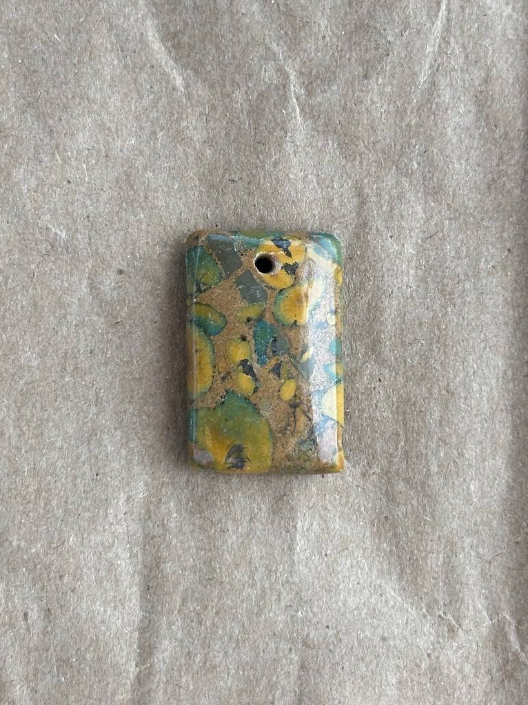 A small rectangular pendant with rounded corners, featuring a marbled pattern of yellow, teal, and brown with a gold leaf accent, displayed on a textured gray background.