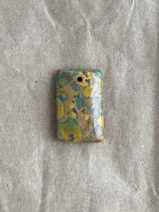 A small rectangular pendant with rounded corners, featuring a marbled pattern of yellow, teal, and brown with a gold leaf accent, displayed on a textured gray background.