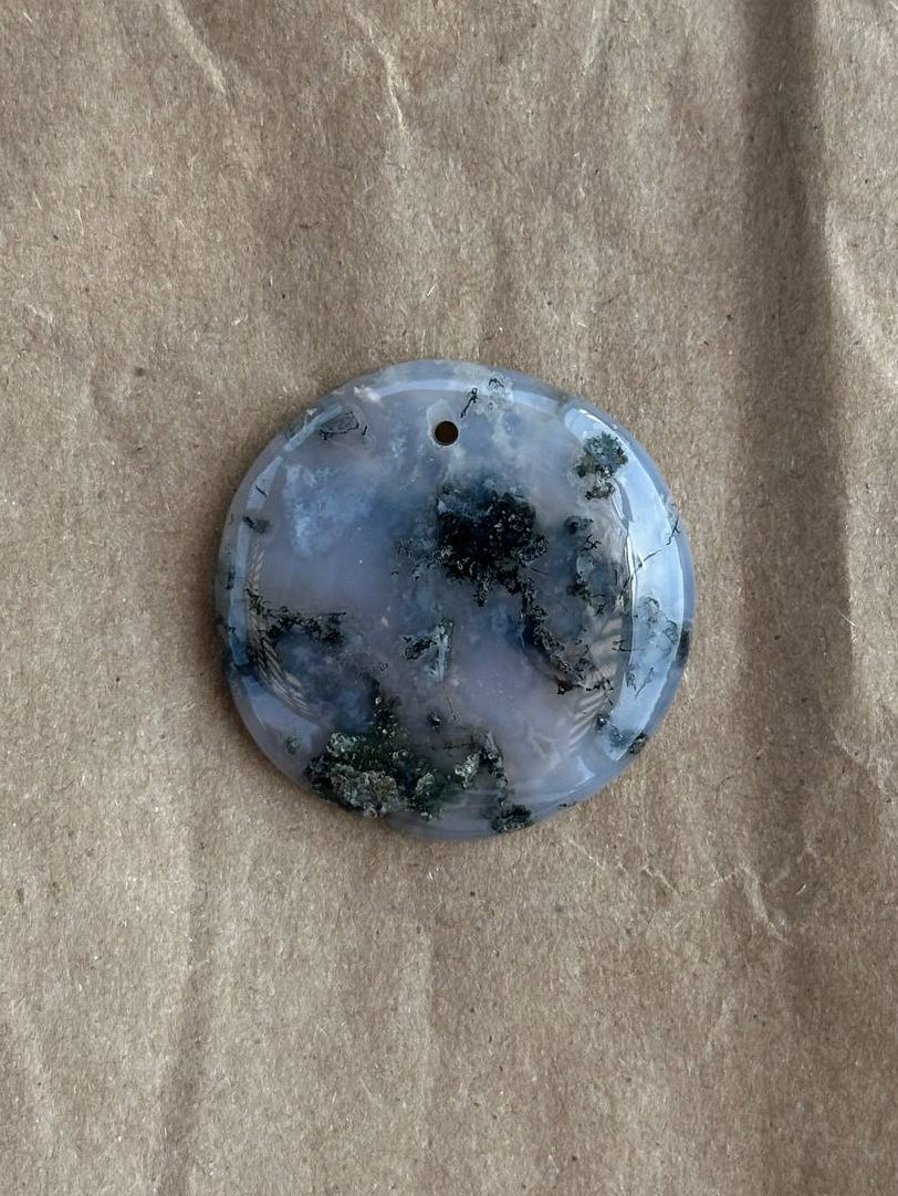 A round polished stone pendant with a small hole at the top, featuring a marbled pattern of light blue, white, and dark green, displayed on a textured brown paper background.