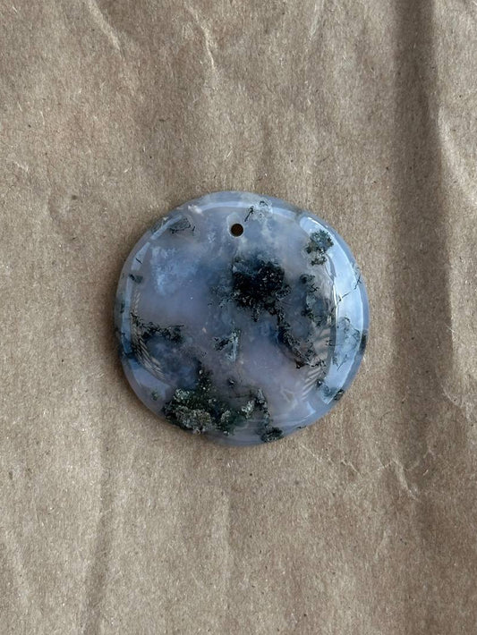 A round polished stone pendant with a small hole at the top, featuring a marbled pattern of light blue, white, and dark green, displayed on a textured brown paper background.
