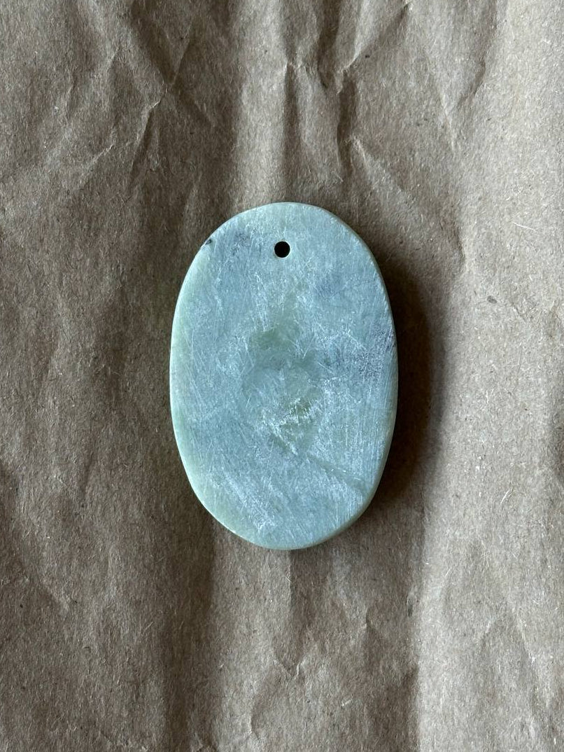 An oval light blue jade pendant with a small hole at the top, resting on a textured brown paper background.