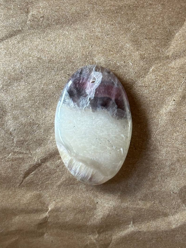 A polished oval gemstone with a natural banded pattern of dark purple and milky white, resting on a textured brown paper background.