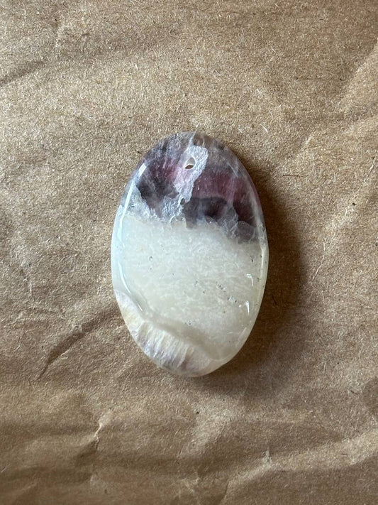 A polished oval gemstone with a natural banded pattern of dark purple and milky white, resting on a textured brown paper background.