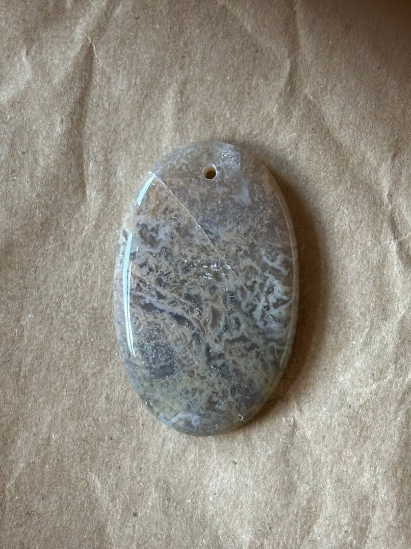 An oval polished stone pendant with a natural marbled pattern in gray, beige, and white, featuring a small hole at the top for hanging, displayed on a textured brown paper background.
